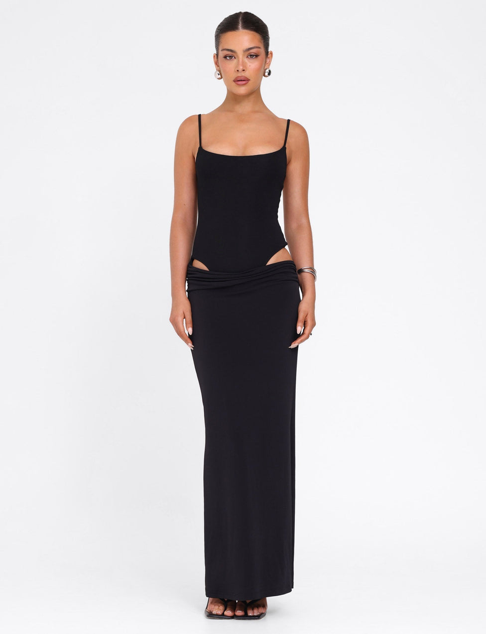 A woman faces forward in the TIGERMIST UMA DRESS - BLACK, a bodycon maxi with adjustable spaghetti straps and cut-out hip details. Her hair is pulled back; she accessorizes with hoop earrings and bracelets, against a plain white background.