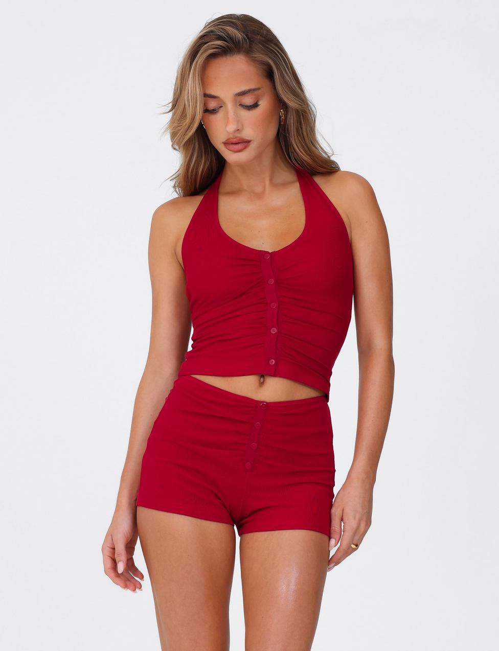 A woman stands against a white background, looking down and to the side, wearing TIGERMIST's ZAFIRA SHORT - RED with a matching sleeveless red crop top featuring front buttons in a body-con fit.