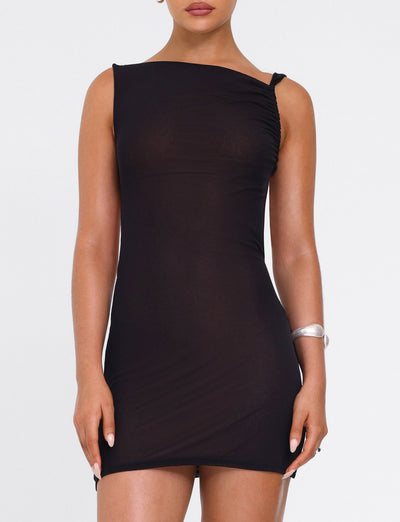 A woman stands against a white background wearing the TIGERMIST FREYA DRESS in black—a fitted, buttery soft mini dress with ruched one-shoulder detail and an adjustable asymmetrical neckline—paired with a silver bracelet on her left wrist.