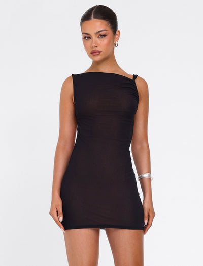 A woman stands against a white background, wearing the TIGERMIST FREYA DRESS in black—a buttery soft mini dress with an adjustable asymmetrical neckline. Her hair is pulled back; she accessorizes with hoop earrings and a silver bracelet.