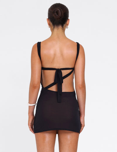 A woman faces away wearing the TIGERMIST FREYA DRESS in black, a buttery soft mini dress with an open back and tie details. Her hair is in a neat bun, accessorized with small hoops and a white bangle on her left wrist.