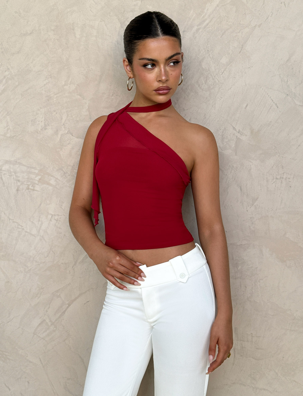 A woman wears the TIGERMIST FIONA TOP in red and white pants, standing against a textured beige wall with one hand on her waist, looking to her left.