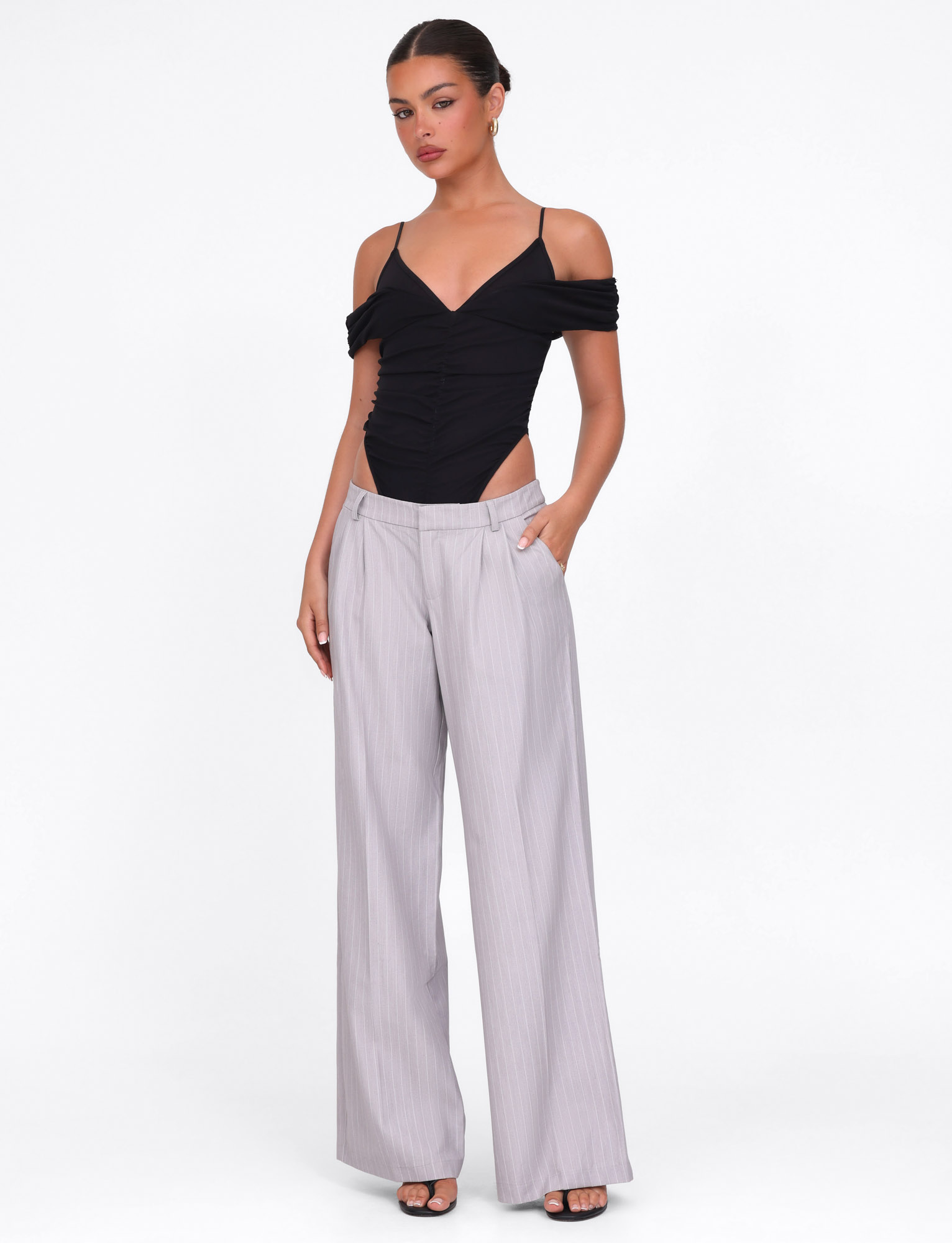 A woman stands against a white background wearing the TIGERMIST AMBERLY SUIT PANT in grey pinstripe with a black off-shoulder top, black open-toe heels, her hair pulled back, and one hand in her pocket.