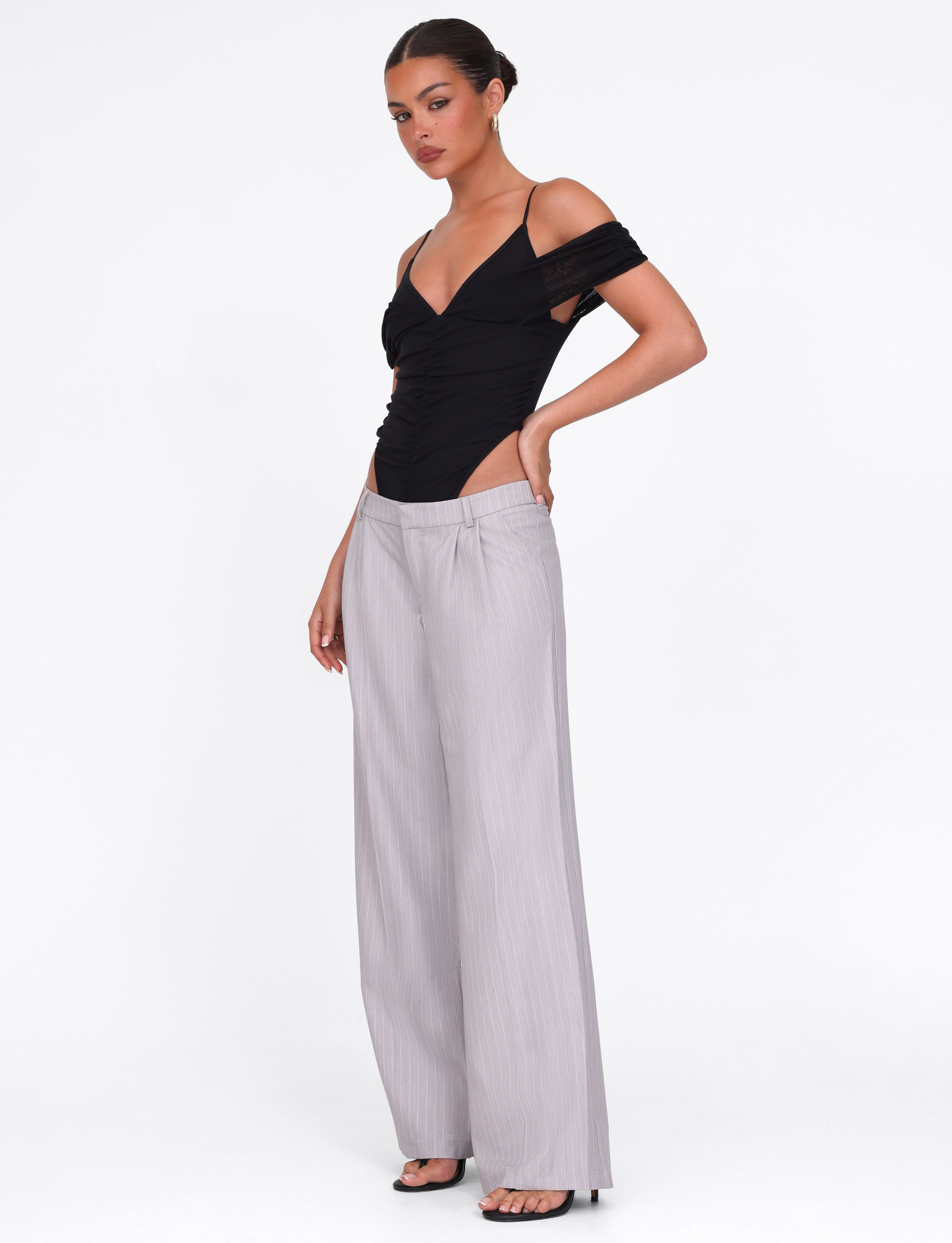 A woman stands against a white background wearing the TIGERMIST AMBERLY SUIT PANT in grey pinstripe, styled with a black off-shoulder top and black open-toe heels. Her hair is pulled back in a sleek bun.