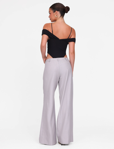 A woman faces away, styled in a black off-shoulder top and TIGERMIST’s AMBERLY SUIT PANT in grey pinstripe. Her hair is in a neat bun as she poses against a plain white background.