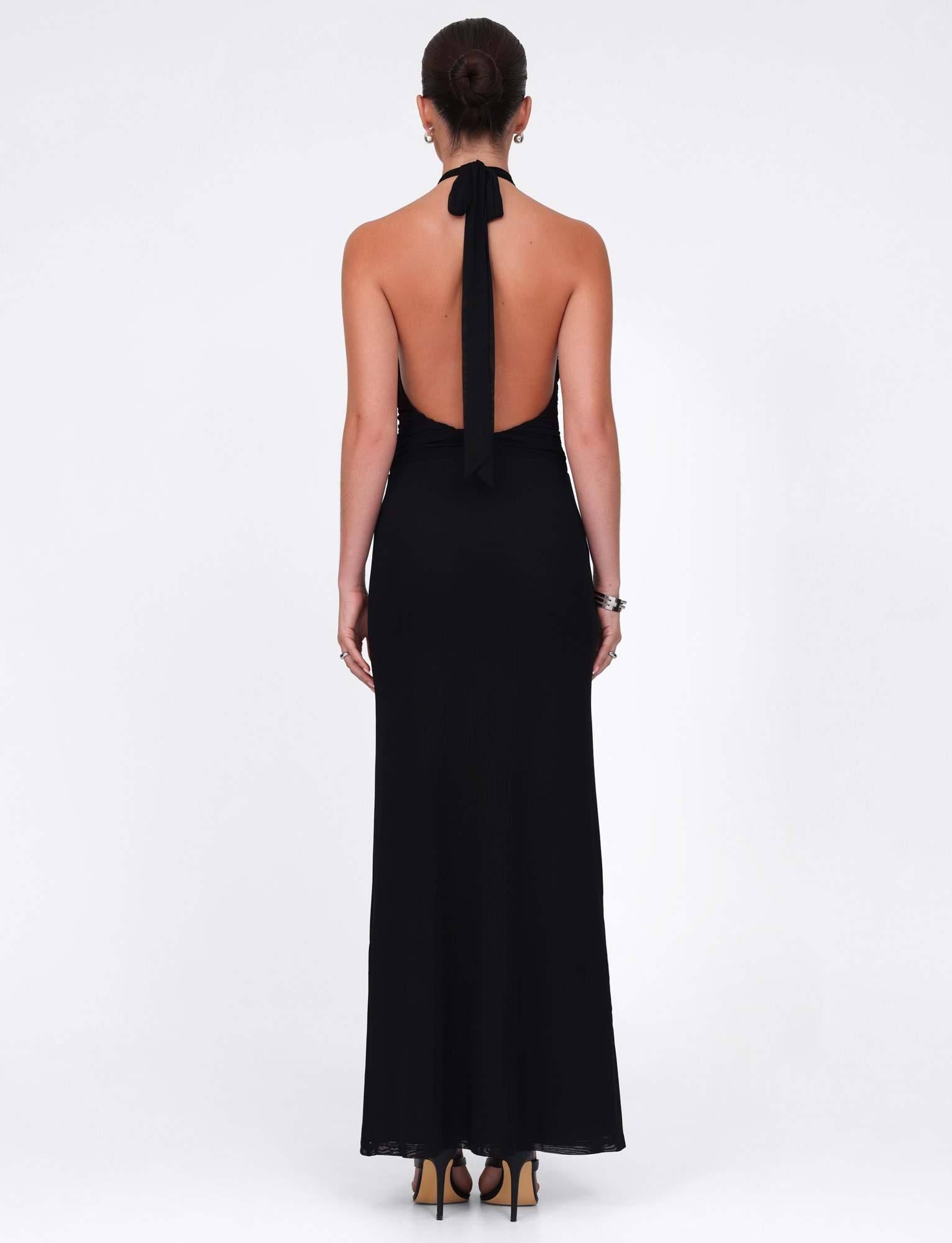 A woman in the TIGERMIST ZARA MAXI DRESS - BLACK, a long halter dress made from high stretch fabric, stands facing away in high heels with her hair in a bun against a plain white background.
