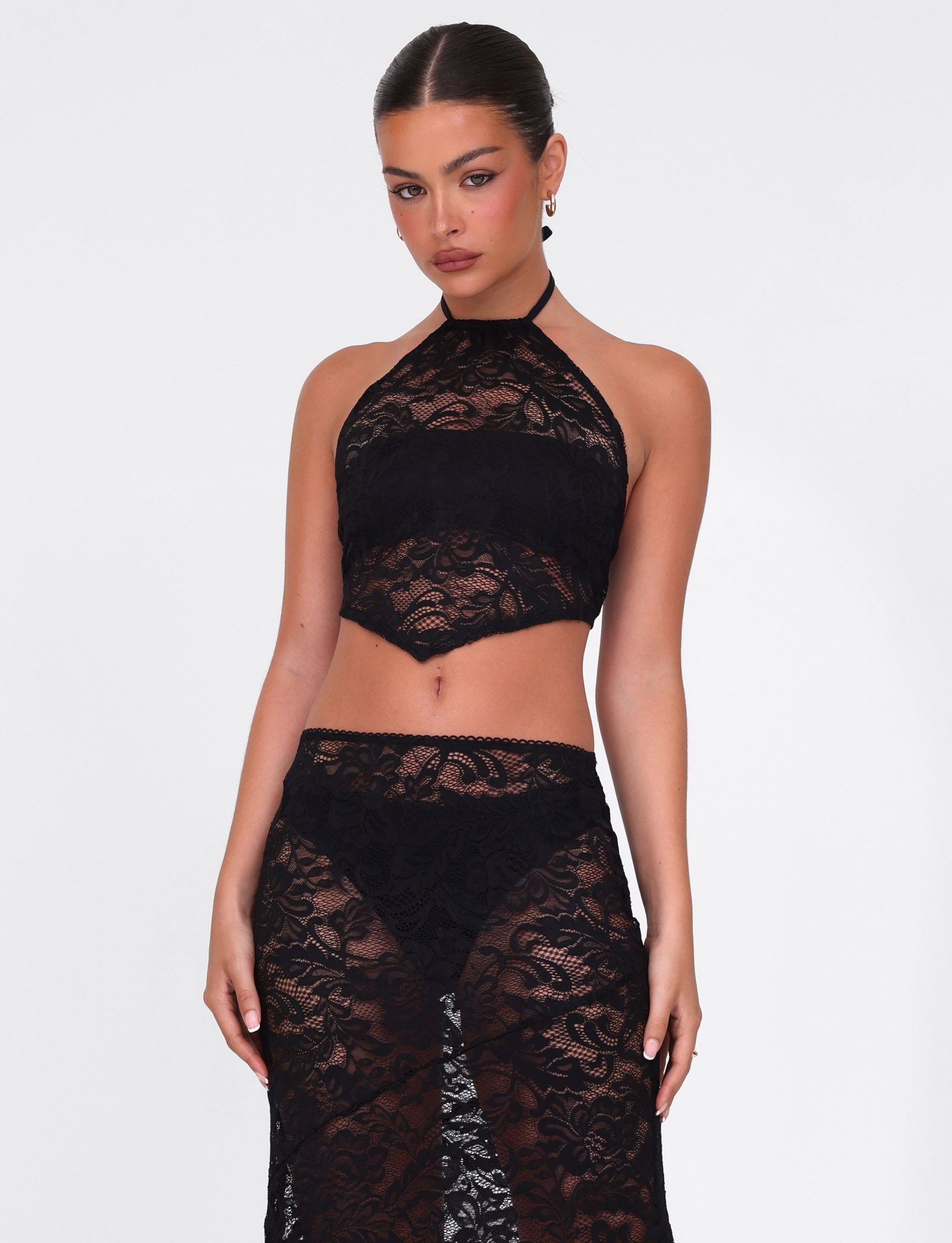 A woman with slicked-back hair wears the TIGERMIST SIYA LACE TOP in black lace, paired with a matching high-waisted skirt, standing against a white background and looking at the camera with a neutral expression.