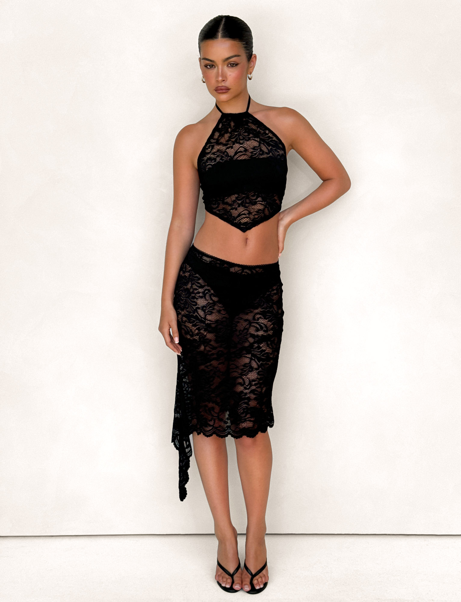 A woman wearing the TIGERMIST SIYA MIDI SKIRT in black lace stands against a light background, pairing the pull-on sheer lace skirt with a matching halter top, black heeled sandals, and minimal jewelry. Her dark hair is pulled back as she poses relaxed with one hand on her hip.