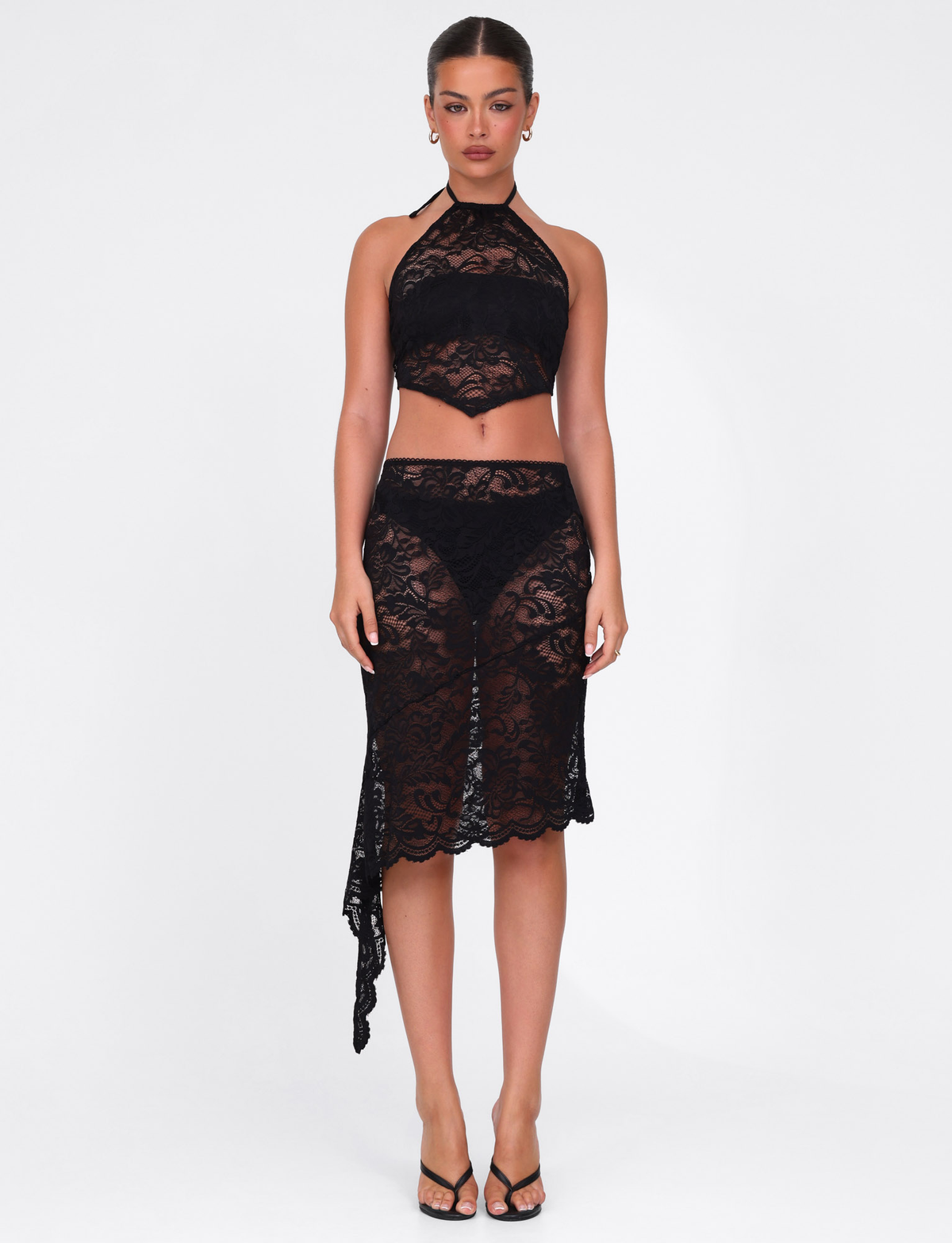 A woman stands against a white background wearing the TIGERMIST SIYA MIDI SKIRT - BLACK LACE and matching black lace halter crop top, styled with black high-heeled sandals. The outfit features easy pull-on construction for effortless style.