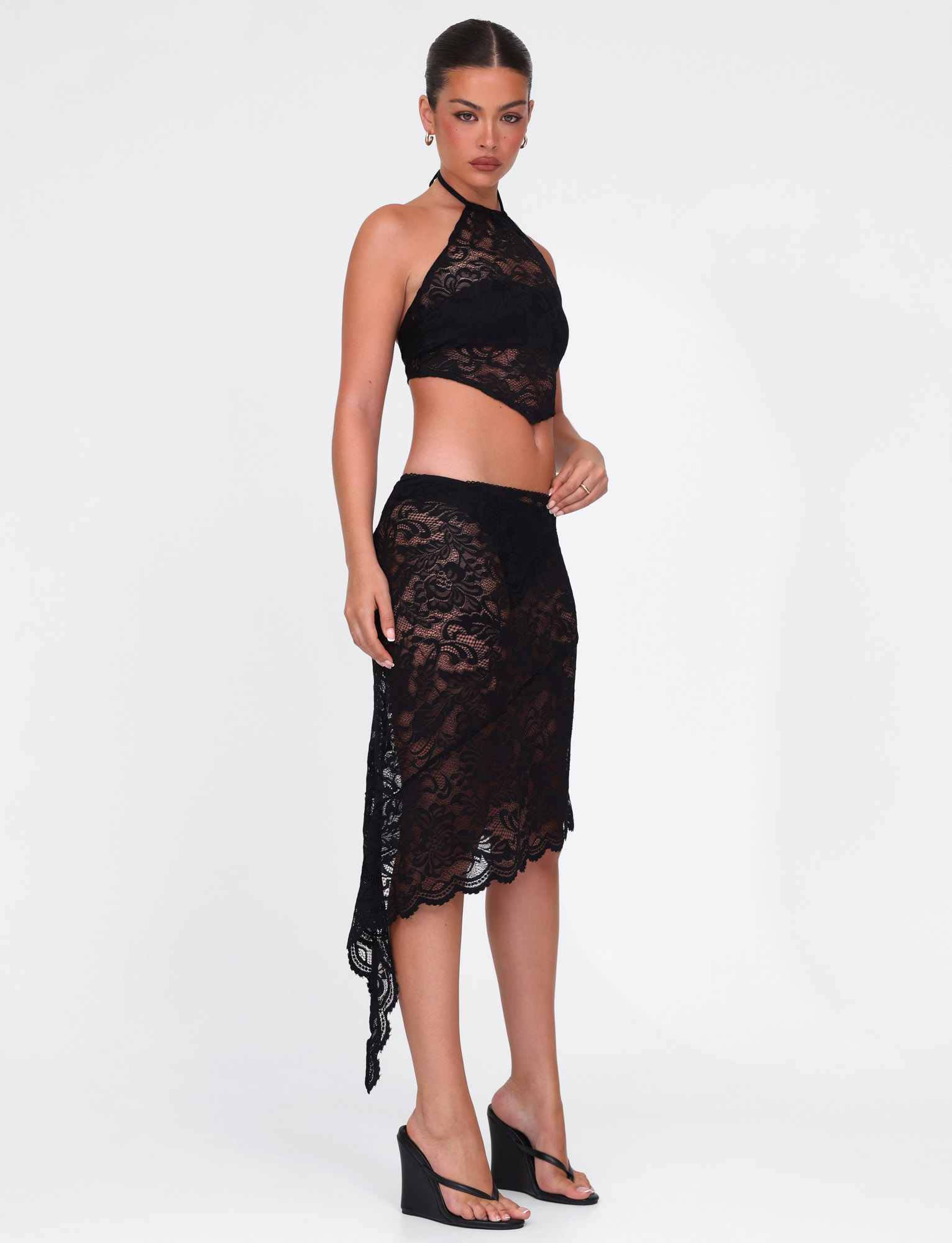 A woman poses against a white background wearing the TIGERMIST SIYA MIDI SKIRT in black lace, featuring a sheer finish and asymmetrical hemline, paired with a matching black lace halter top and black wedge sandals. Her hair is tied back.