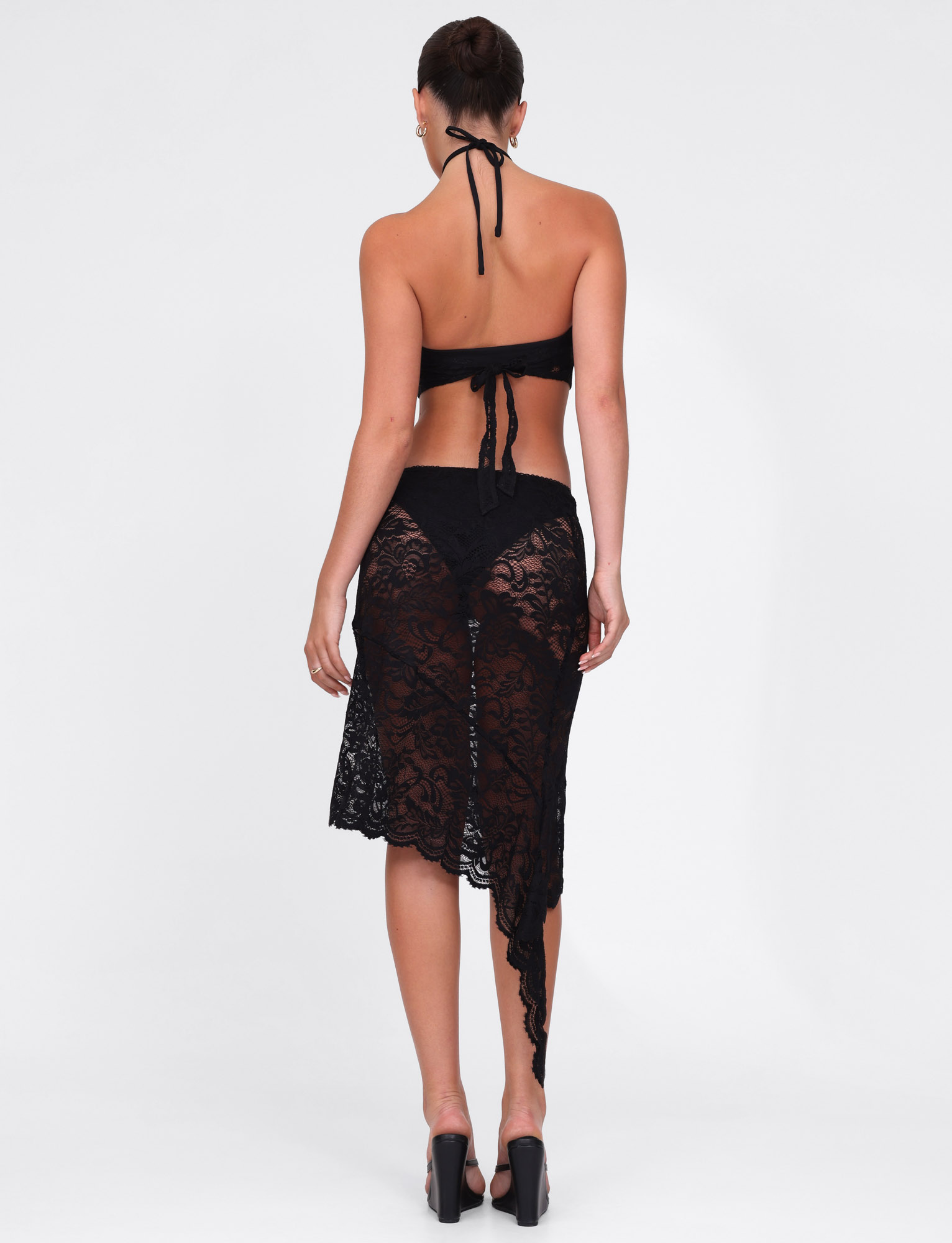 A woman models the TIGERMIST SIYA MIDI SKIRT in black lace with an asymmetrical hemline, styled with a black halter top and high heels, standing against a plain white backdrop.