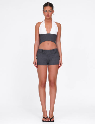 A woman faces forward wearing the TIGERMIST LOTUS TOP in cream and high-waisted grey shorts, paired with hoop earrings and black heeled sandals. Her hair is pulled back, set against a plain white background.