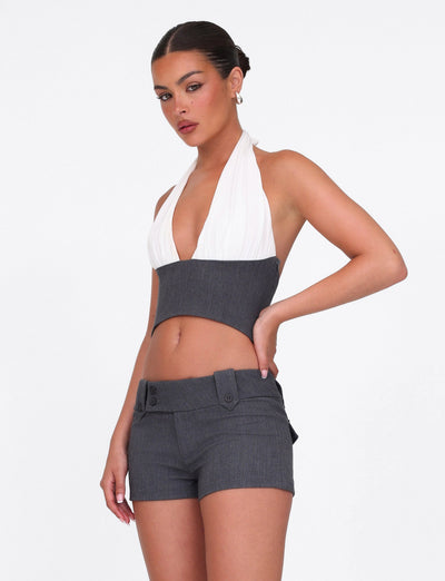A woman poses confidently in the TIGERMIST LOTUS TOP - CREAM : GREY, a sleeveless, cropped halter with a plunging neckline, paired with high-waisted gray shorts featuring wide belt loops and button details against a white background.