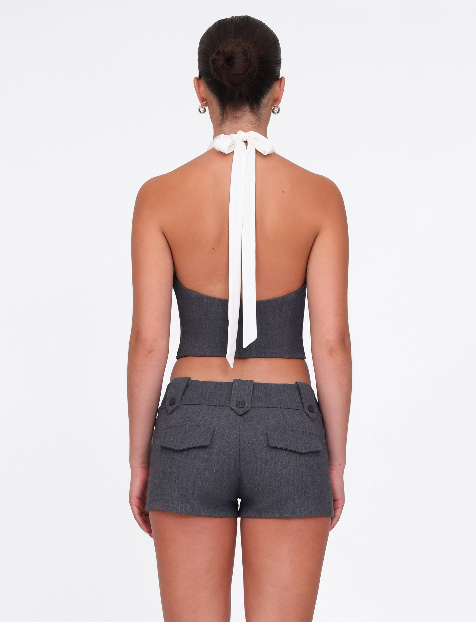 A woman with her hair in a bun is seen from behind wearing the TIGERMIST LOTUS TOP - CREAM : GREY, a dark gray cropped halter top with a white neck tie and matching high-waisted shorts, both in comfy stretch fabric. Background is plain white.