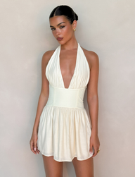 A woman stands against a light background wearing the TIGERMIST CALISTA MINI DRESS - YELLOW : BUTTER, featuring a halter neck, fitted waist panel, deep V-neck, and flared skirt. Her hair is styled back, paired with earrings and a ring.