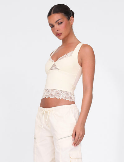 A person wearing the TIGERMIST Dione Lace Top in Soft Butter and light cargo pants poses against a white background. With tied-back hair, they look to the side with a neutral expression, embodying casual elegance.