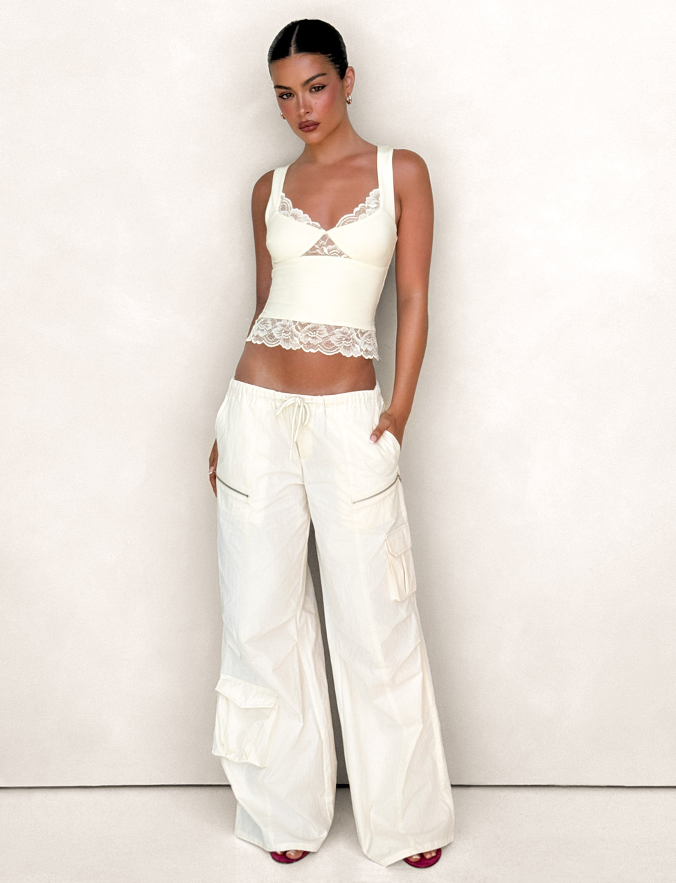 A woman poses confidently with slicked-back dark hair, wearing the TIGERMIST HERMIONE PANT in Soft Butter Yellow—stylish wide-leg cargo pants with a drawstring waist and side pockets—paired with a fitted ivory lace-trim tank top.