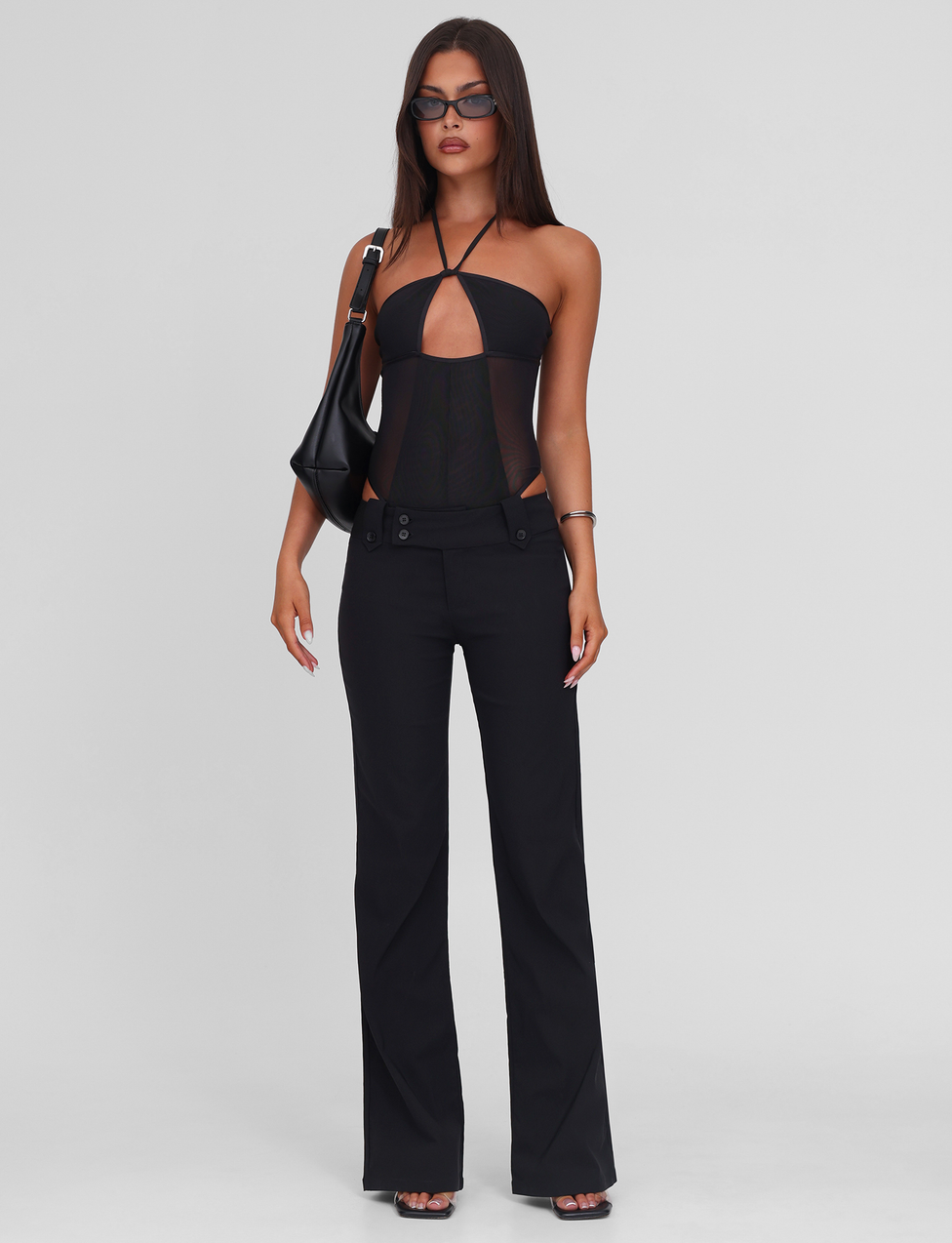 A woman wearing TIGERMIST's MELANIA BODYSUIT in black with sunglasses, high-waisted flared stretch pants, black open-toe heels, and a black adjustable-strap shoulder bag stands against a plain light background.