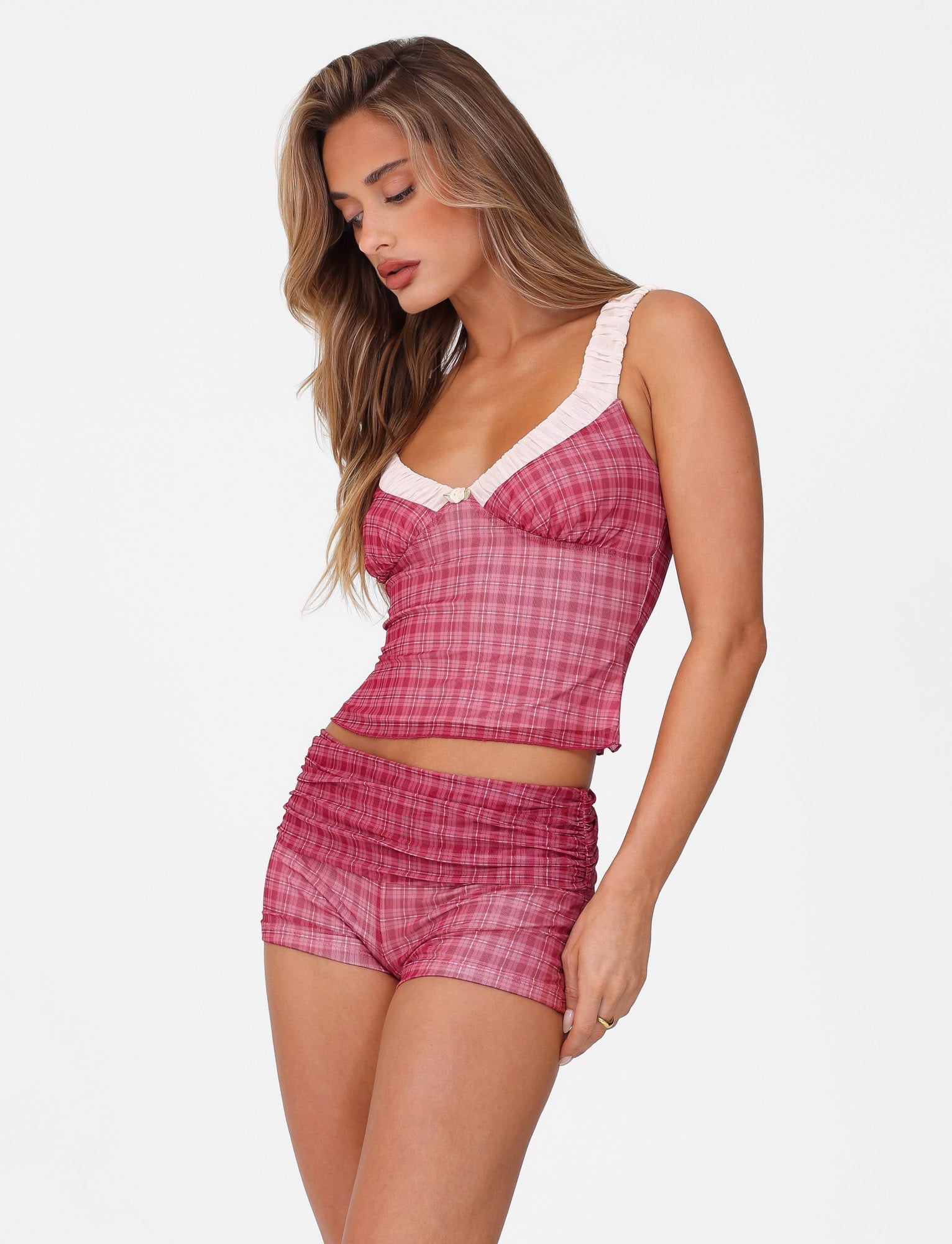 A woman models the TIGERMIST AMARAN SHORT - RED : RED CHECK set. With a bodycon fit and high-waisted shorts, her pink plaid ensemble contours elegantly against a white backdrop. Her long, wavy hair cascades down as she looks downward, enhancing the outfit's high stretch fabric appeal.
