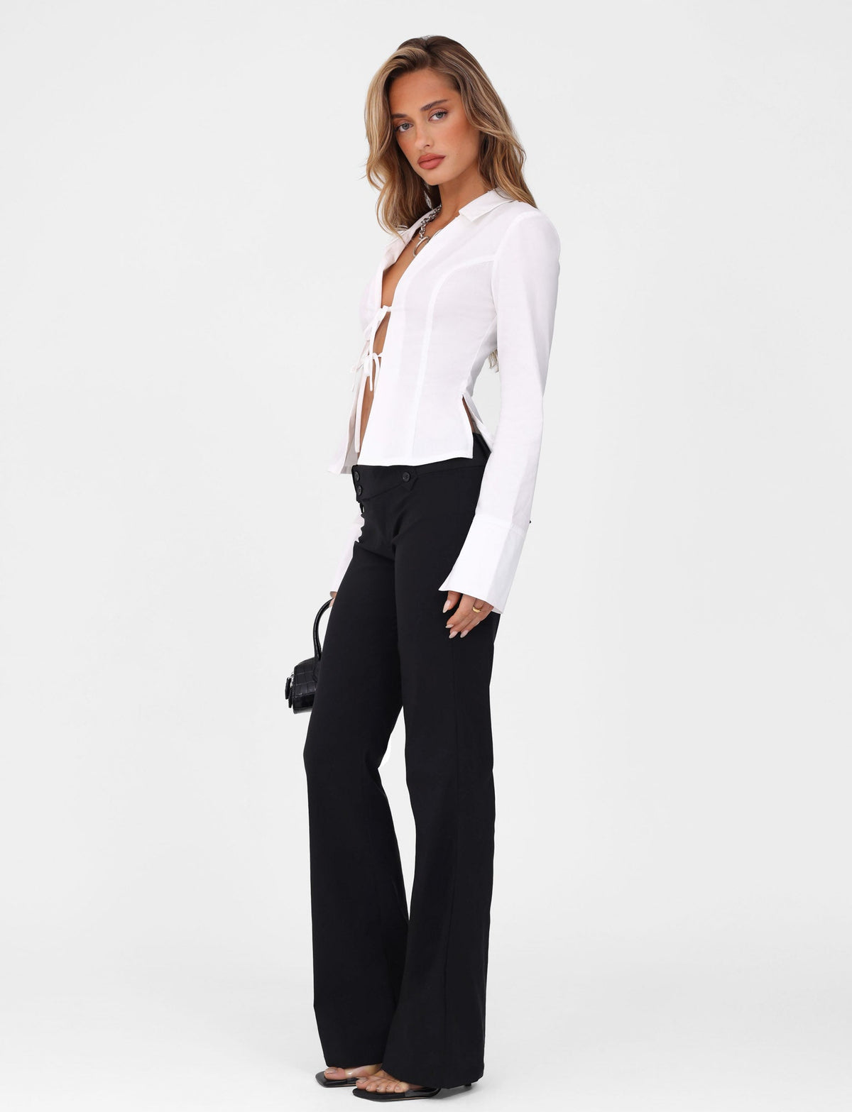 A person with long hair poses against a white background wearing TIGERMIST's BRIELLE SHIRT - WHITE : IVORY and black wide-legged pants. They hold a small black bag and wear strappy black sandals.