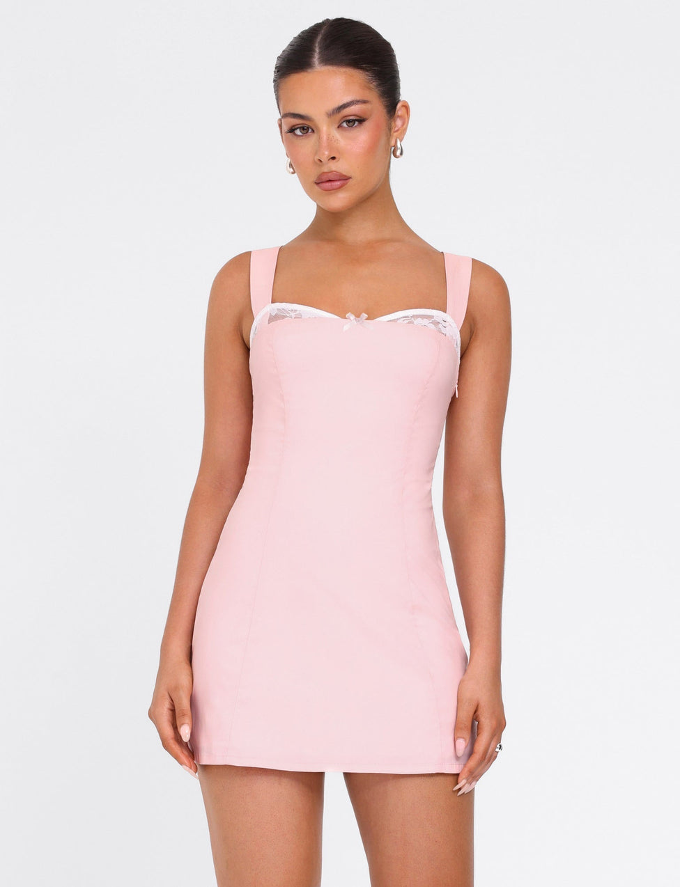 A woman models the TIGERMIST COLSON MINI DRESS in baby pink, featuring wide straps and a lace-trimmed neckline, as she stands against a plain white background and looks at the camera.