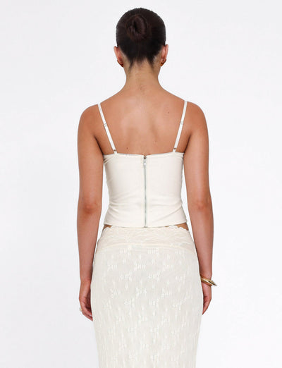 A woman faces away from the camera in the TIGERMIST ANTONELLA TOP—bone cream color with adjustable straps and a visible back zipper—paired with a sheer lace-patterned skirt; her dark hair is styled in a neat bun.