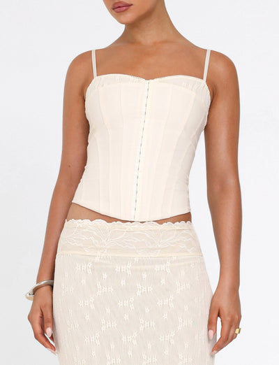A woman wears the TIGERMIST ANTONELLA TOP in cream, featuring adjustable straps and vertical seams, paired with a matching sheer lace skirt. Her face is partially out of frame as she stands before a plain white background.