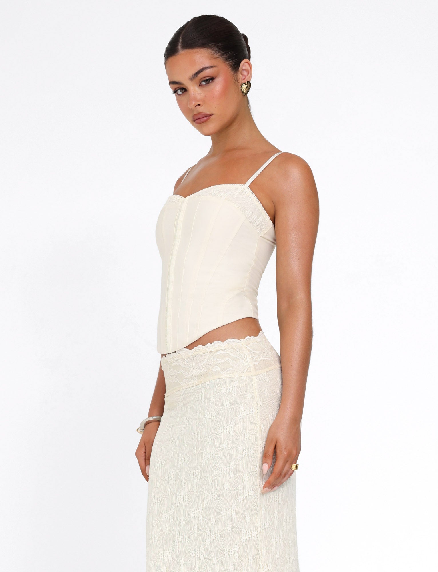 A woman wears the TIGERMIST ANTONELLA TOP in white/cream with adjustable straps and a matching lace skirt, standing against a plain white background and gazing over her shoulder with a neutral expression.