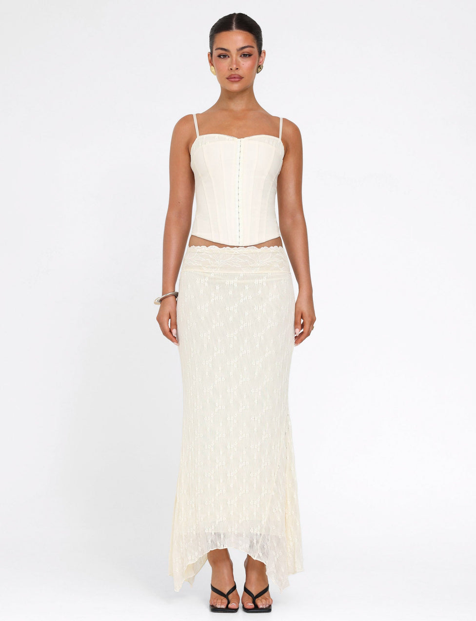 A woman stands against a plain white background, wearing the TIGERMIST ANTONELLA MIDI SKIRT in lace cream with a handkerchief hem and a cream corset top. Her hair is pulled back as she looks directly at the camera.