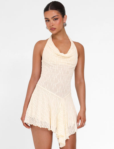 A woman wears the TIGERMIST FIORELLA MINI DRESS in cream lace with a halter neck and asymmetrical hem, standing against a white background and looking at the camera.
