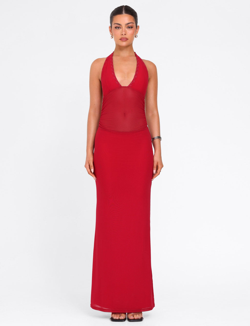 A woman stands facing forward in the TIGERMIST BRESILY MAXI DRESS - RED, a sleeveless, floor-length dress with halter straps and a sheer midsection. Her hair is pulled back; she wears black sandals and a silver bracelet against a plain white background.