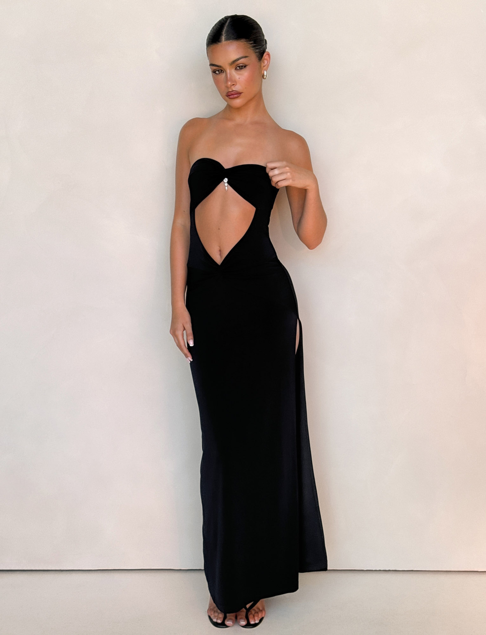 A woman, hair styled back and wearing subtle silver jewelry, poses against a plain wall in the TIGERMIST GRAZIA DRESS - BLACK: a stretch bandeau maxi with front cutout and thigh-high slit, paired with open-toe heels.