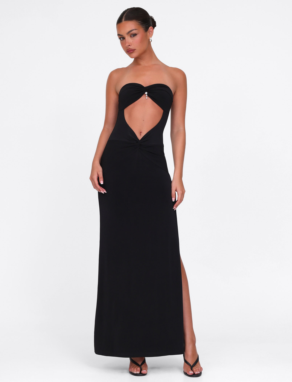 A woman models the TIGERMIST GRAZIA DRESS - BLACK, a strapless bandeau maxi in stretch fabric with a front cutout, standing with one leg forward and hands on hips against a plain white background.