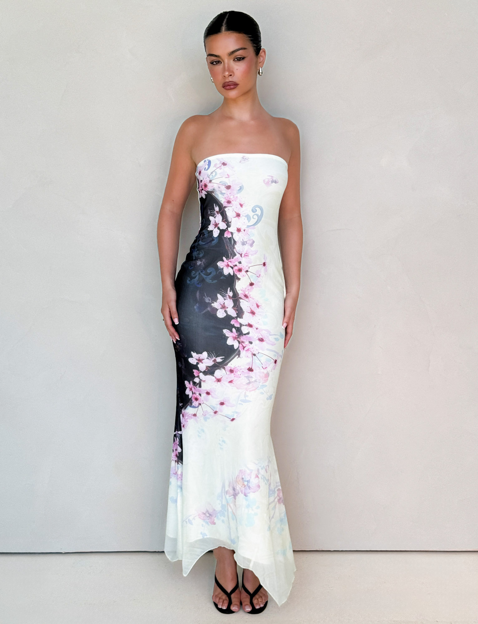 A woman stands against a light wall in the TIGERMIST LILLIAN MAXI DRESS - MULTI: FLORAL: CHERRY BLOSSUM, featuring black, white, and pink floral mesh. Her sleek hair is pulled back and she pairs it with open-toe black heels.