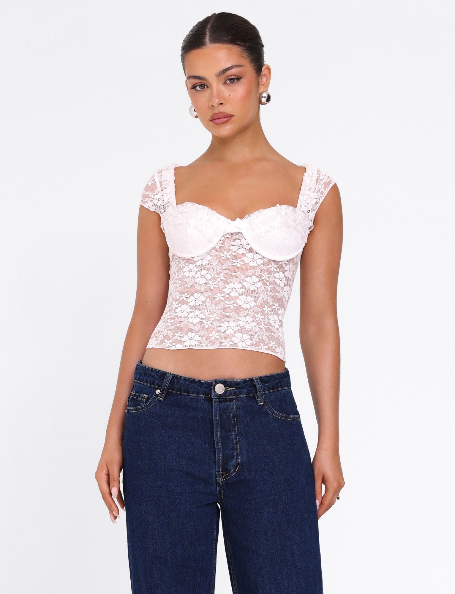 A woman stands against a plain white background wearing the TIGERMIST NYRA TOP in white, a fitted semi-sheer lace bustier with cap sleeves, paired with high-waisted dark blue jeans. Her hair is pulled back and she accessorizes with pearl earrings.