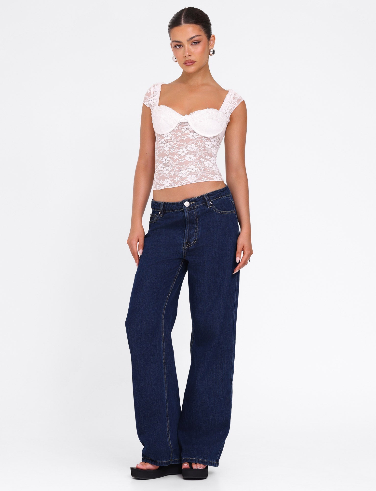 A woman models the TIGERMIST NYRA TOP - WHITE, a white lace semi-sheer bustier with cap sleeves and sweetheart neckline, paired with dark blue wide-leg jeans, black platform sandals, and hoop earrings against a white background.