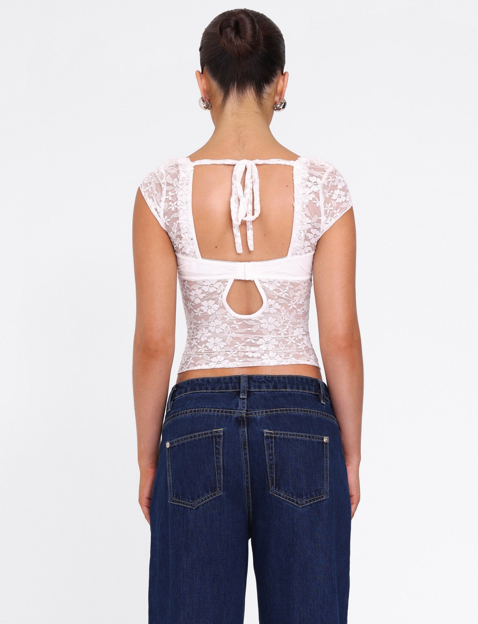 A woman faces away, wearing the TIGERMIST NYRA TOP in white—a semi-sheer lace top with open back and tie detail—paired with high-waisted dark blue jeans. Her hair is styled in a neat bun.