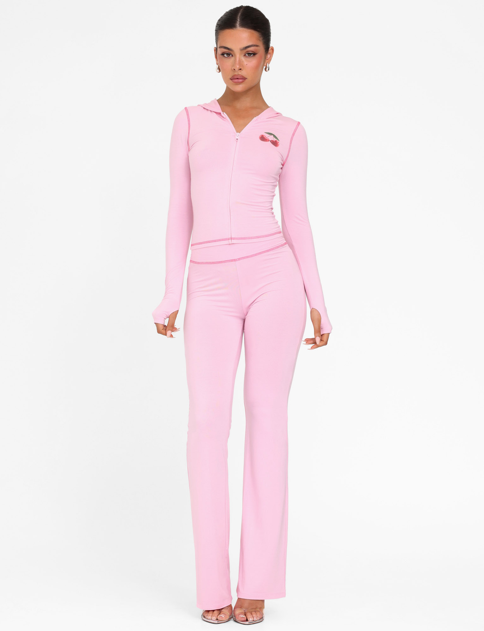 A woman faces forward in a fitted TIGERMIST BLAYZE HOODIE in soft pink with a chest patch and matching high-waisted flared pants, styled with open-toe heels against a plain white background.