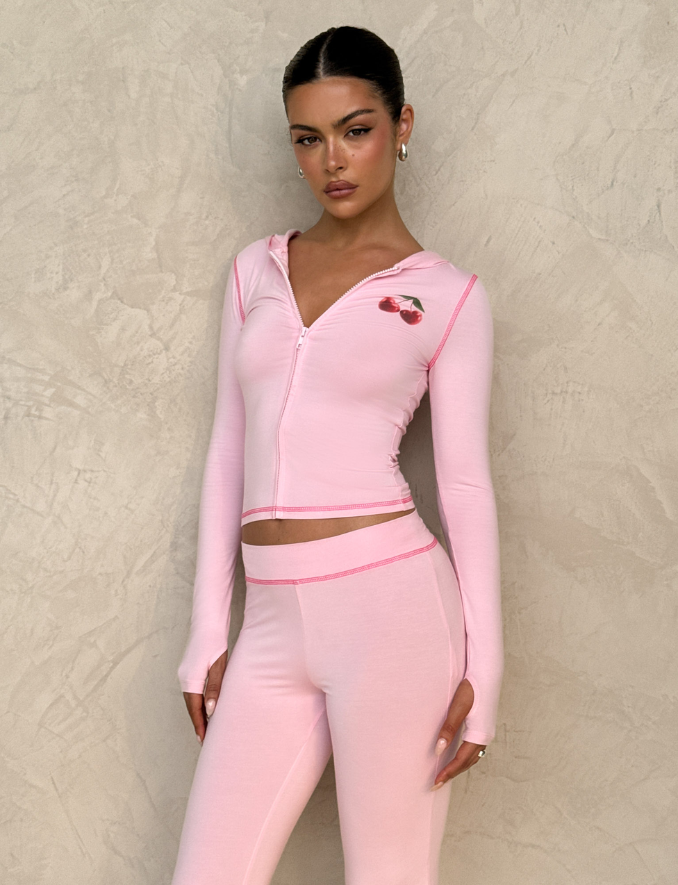 A woman wears the TIGERMIST BLAYZE HOODIE in soft pink, styled with matching leggings, as she stands against a textured beige wall and gazes confidently at the camera.