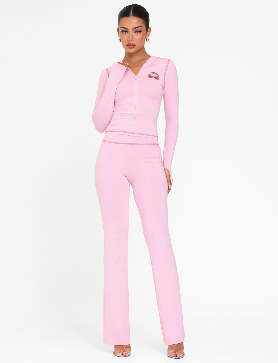 A woman poses against a white background wearing TIGERMIST’s BLAYZE PANT in soft pink with a matching fitted hoodie and clear open-toe heels, hand near her chin as she gazes at the camera.