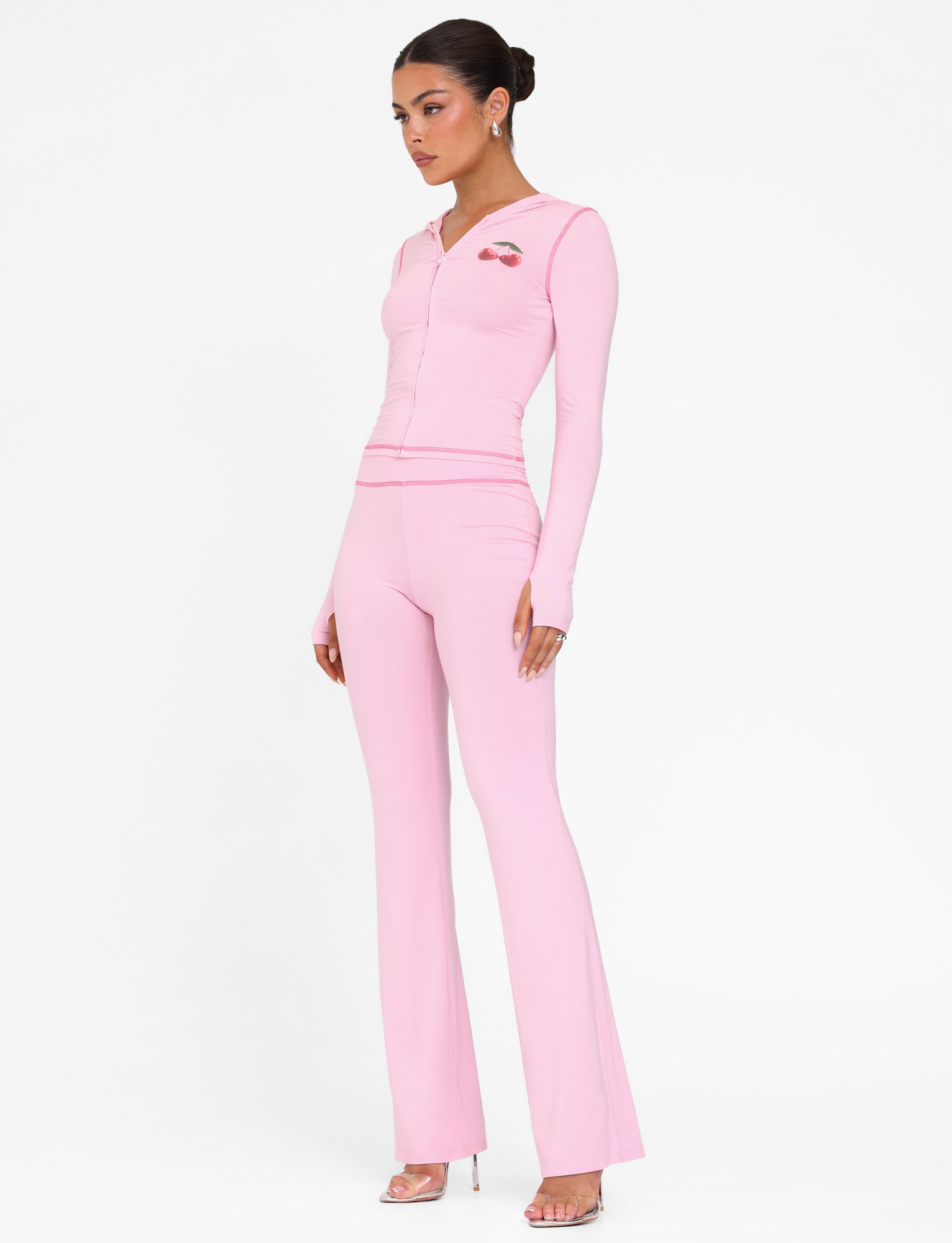 A woman models the TIGERMIST BLAYZE PANT - PINK in soft pink against a white background, pairing the flared pants with a matching fitted zip-up top and open-toe heels. Her hair is in a neat updo as she looks off to the side.