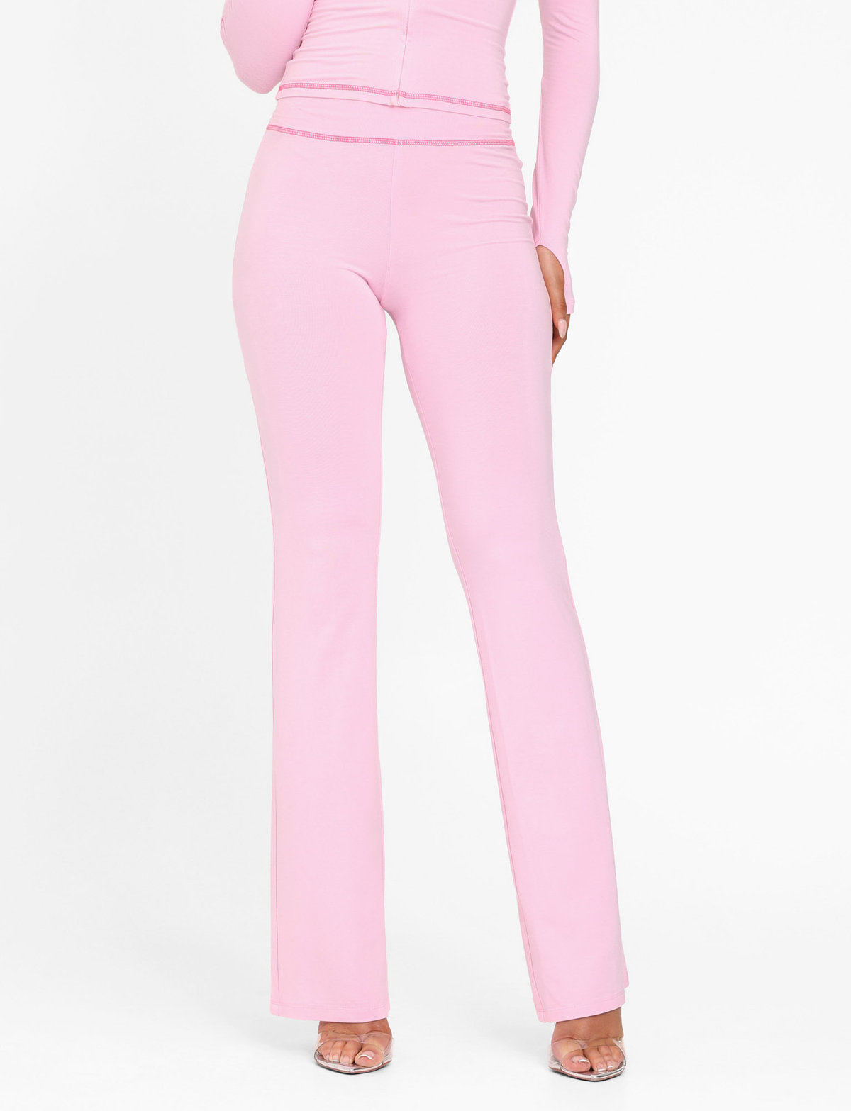 Wearing TIGERMIST’s BLAYZE PANT in soft pink, paired with a matching long-sleeve top and clear heels, this person poses against a plain white background.