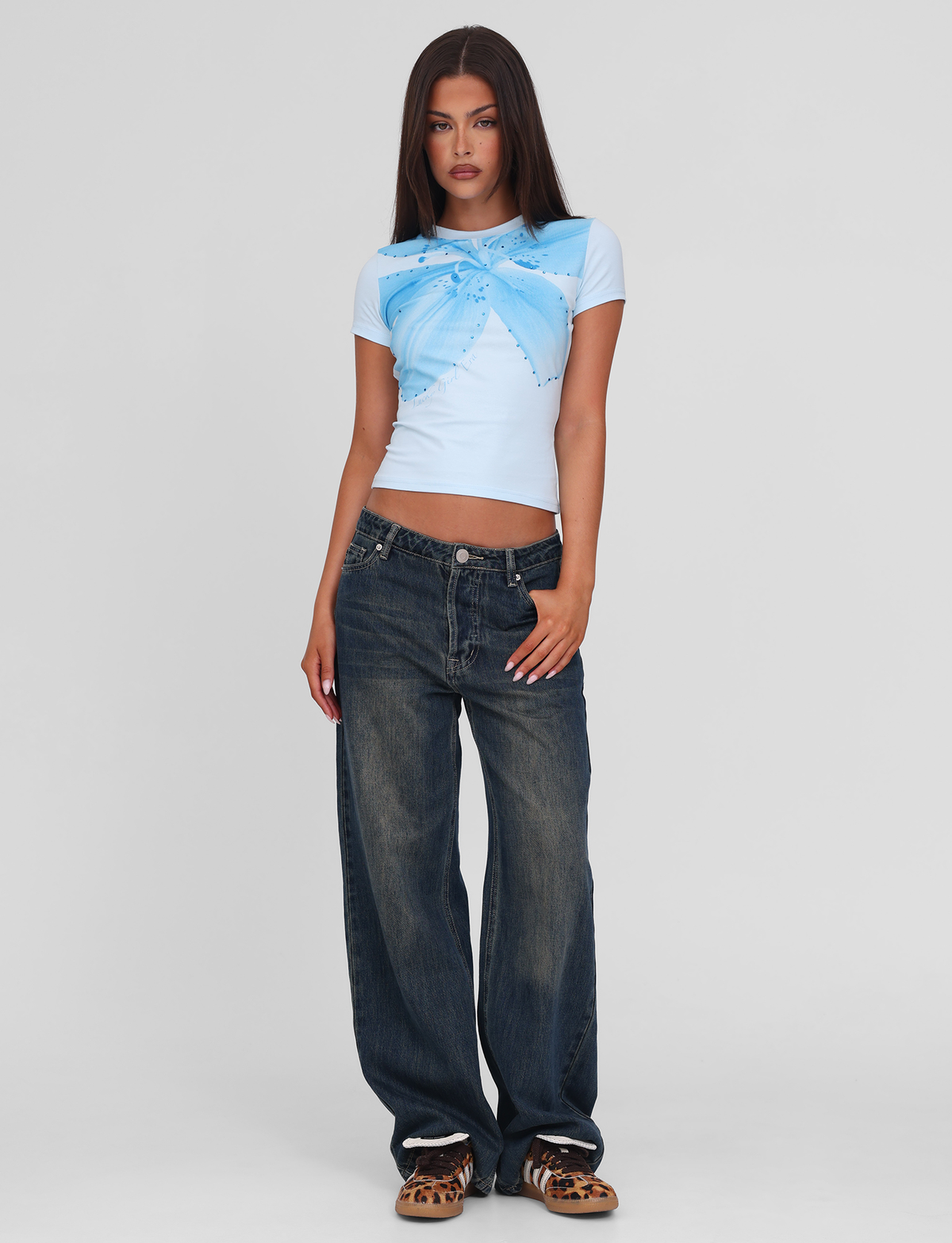 A woman with straight, long brown hair and a neutral expression poses against a plain background wearing the TIGERMIST SAPHIRA TEE - BLUE, featuring a butterfly and floral graphic, paired with loose dark blue jeans and leopard-print shoes.