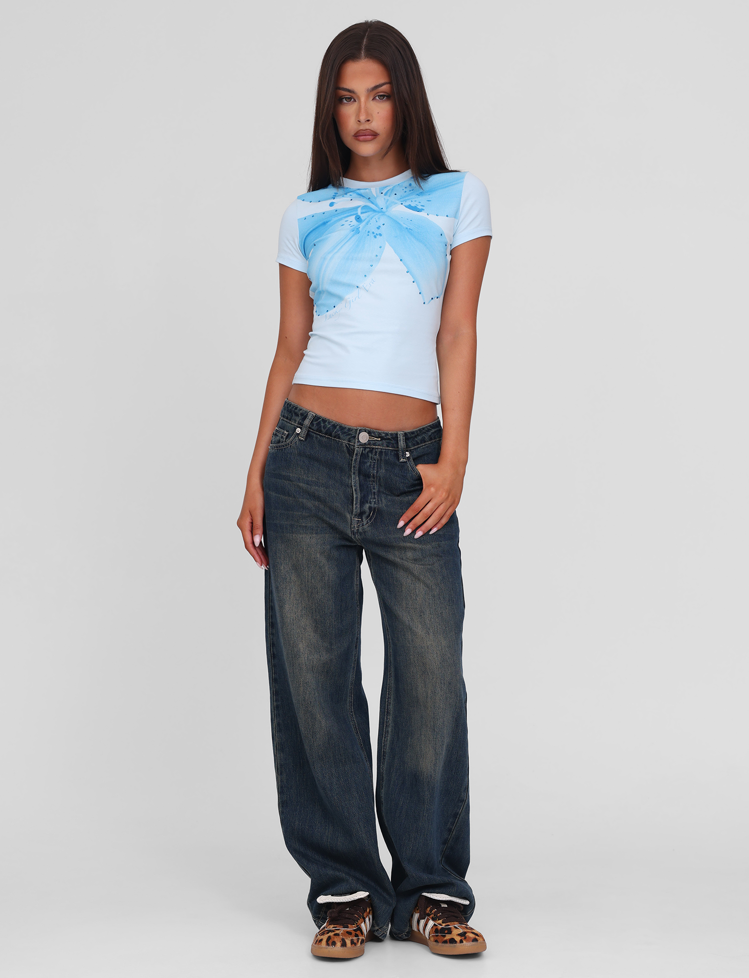 A woman with straight, long brown hair and a neutral expression poses against a plain background wearing the TIGERMIST SAPHIRA TEE - BLUE, featuring a butterfly and floral graphic, paired with loose dark blue jeans and leopard-print shoes.
