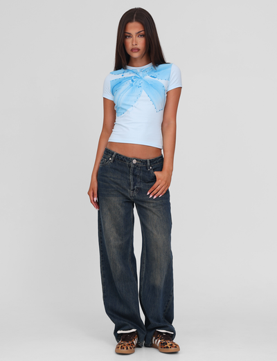 A woman with straight, long brown hair and a neutral expression poses against a plain background wearing the TIGERMIST SAPHIRA TEE - BLUE, featuring a butterfly and floral graphic, paired with loose dark blue jeans and leopard-print shoes.