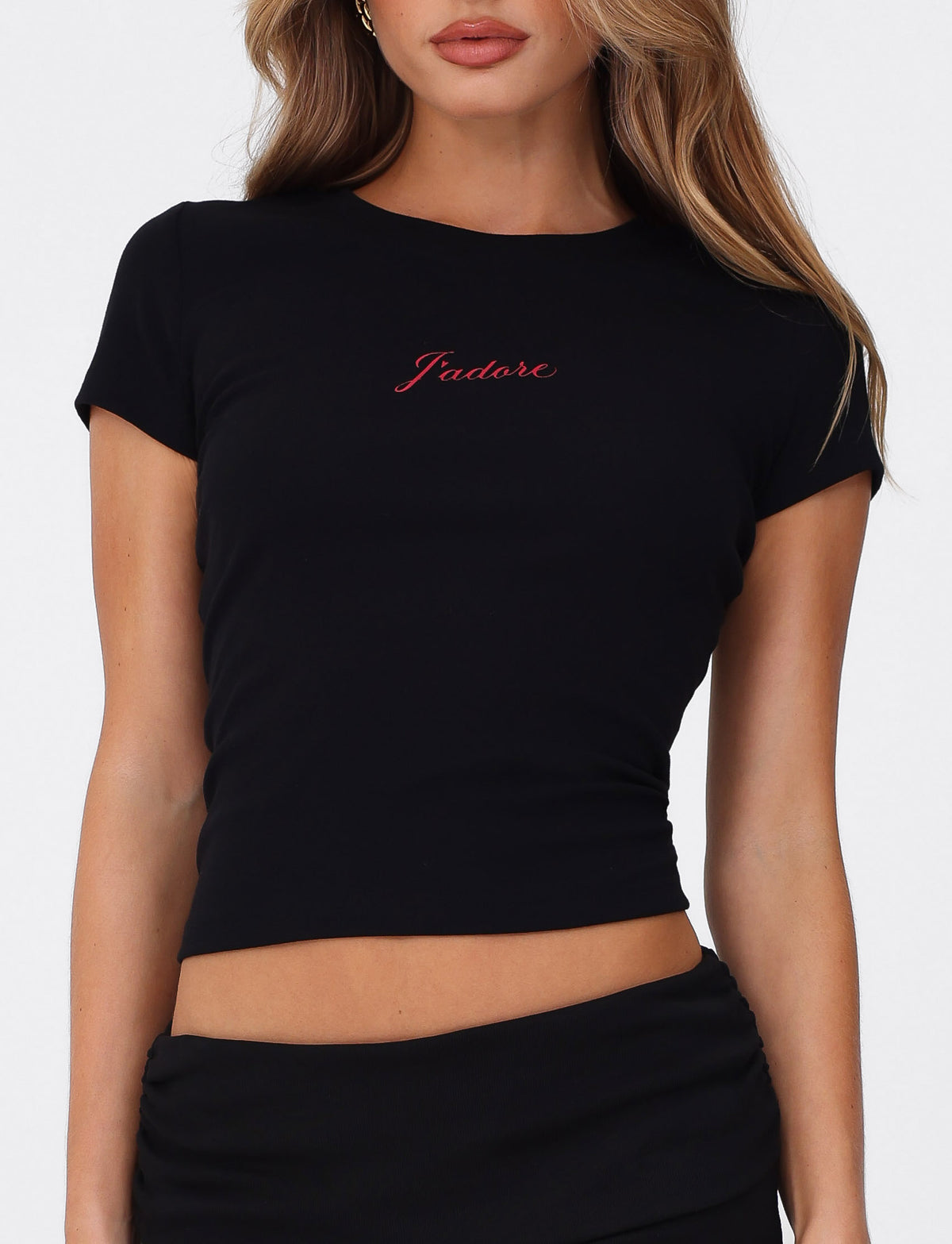 A woman wears the TIGERMIST SAPHIRA TEE in black—a fitted, high-stretch crop top with a red J'Adore graphic—paired with a black skirt and posed against a plain white background.