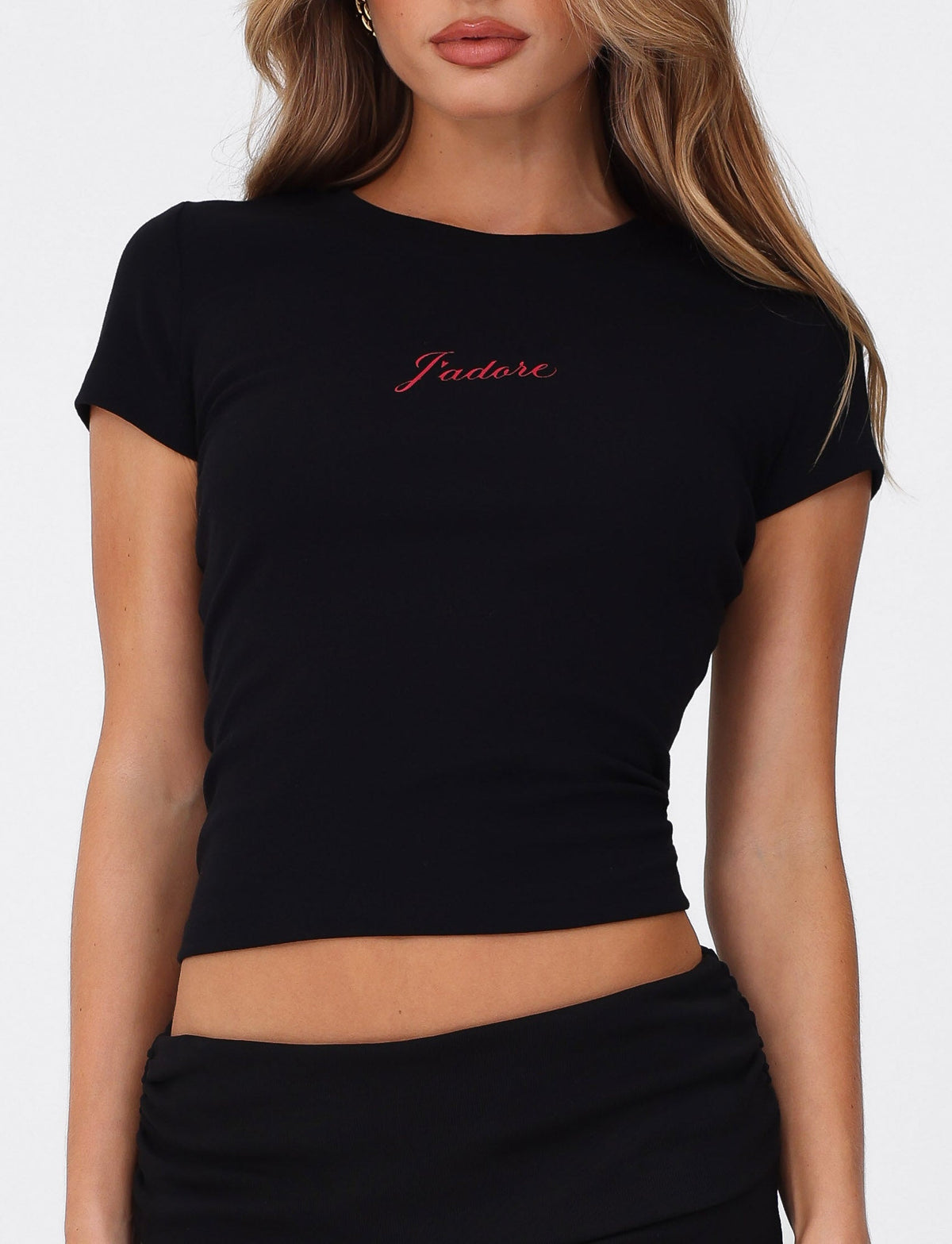 A woman wears the TIGERMIST SAPHIRA TEE in black—a fitted, high-stretch crop top with a red J'Adore graphic—paired with a black skirt and posed against a plain white background.