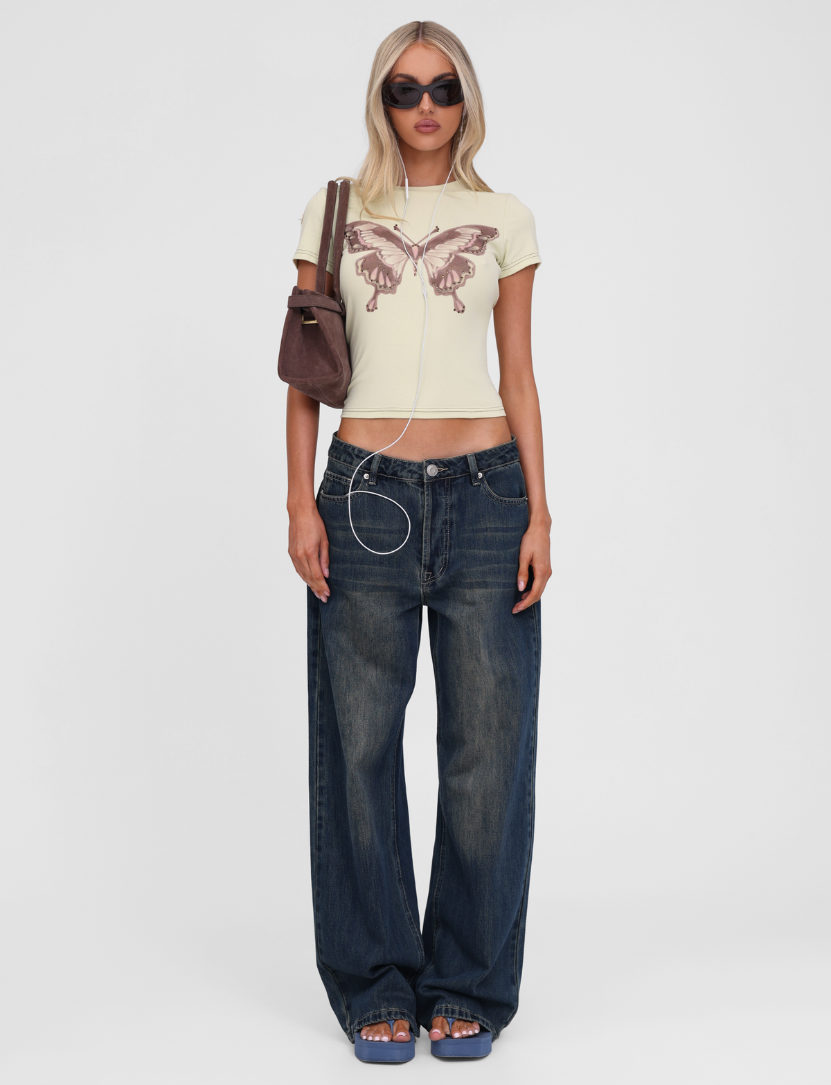 A woman stands facing forward wearing TIGERMIST's SAPHIRA TEE in butterscotch, dark blue jeans, sandals, sunglasses, and a brown shoulder bag. She also has earphones with a visible cord.