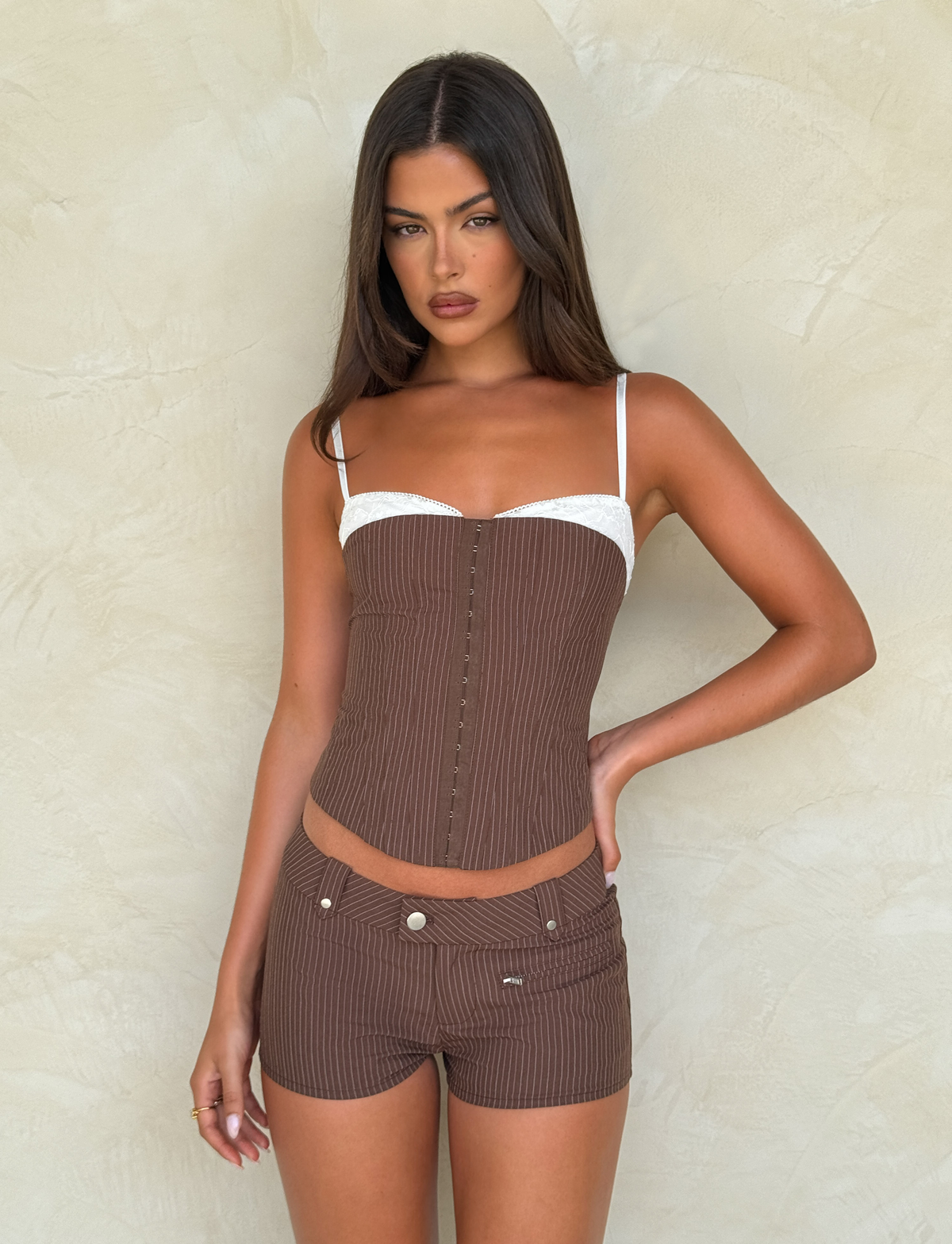 A woman with long brown hair wears the TIGERMIST ANTONELLA CORSET 2.0 in chocolate pinstripe with adjustable straps and matching shorts, standing against a beige textured wall.