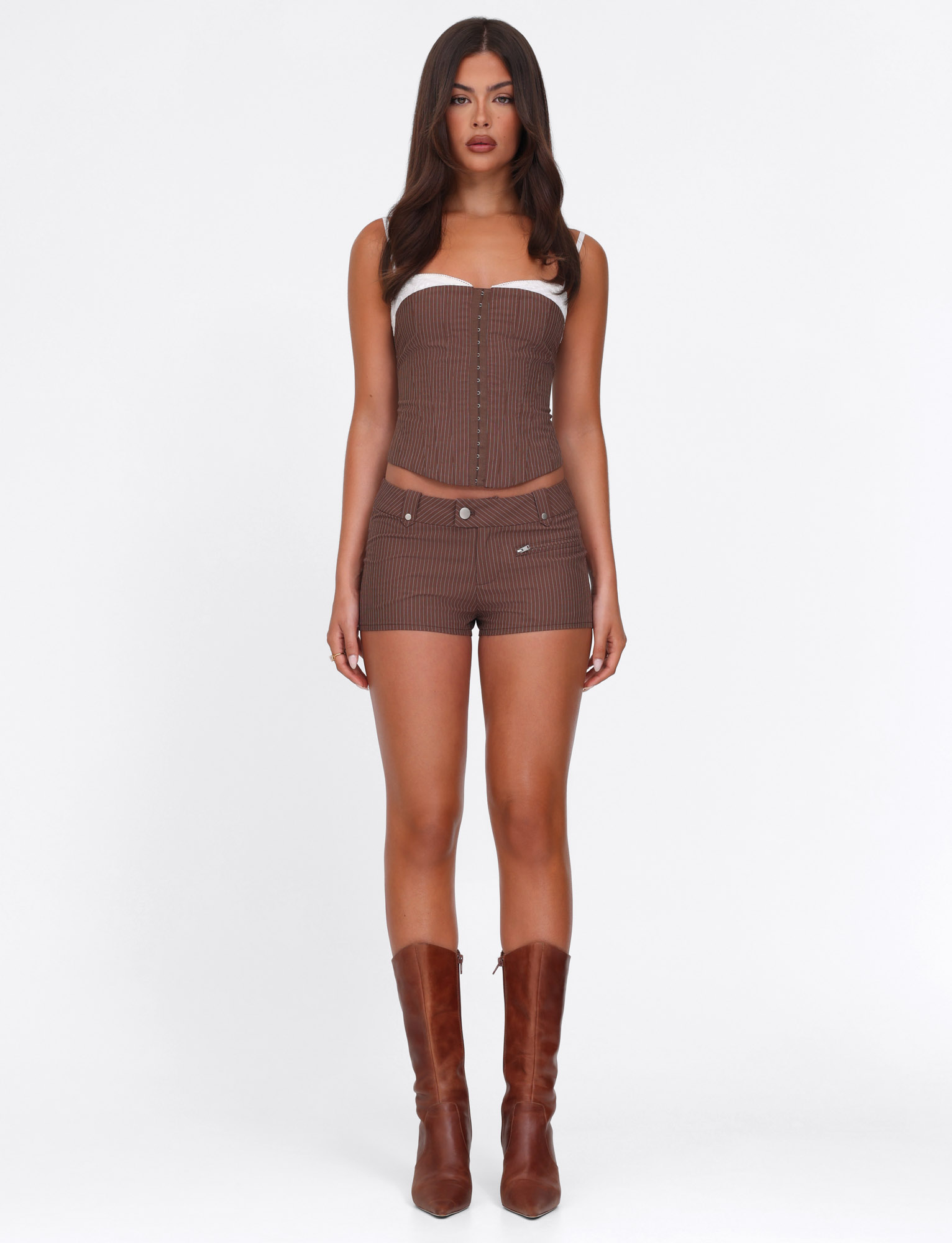 A woman poses against a white background in TIGERMIST’s ANTONELLA SHORT - BROWN : CHOCOLATE : CHOCOLATE PINSTRIPE, styled with a brown strapless pinstripe corset top and knee-high boots. Her hair falls in loose waves.